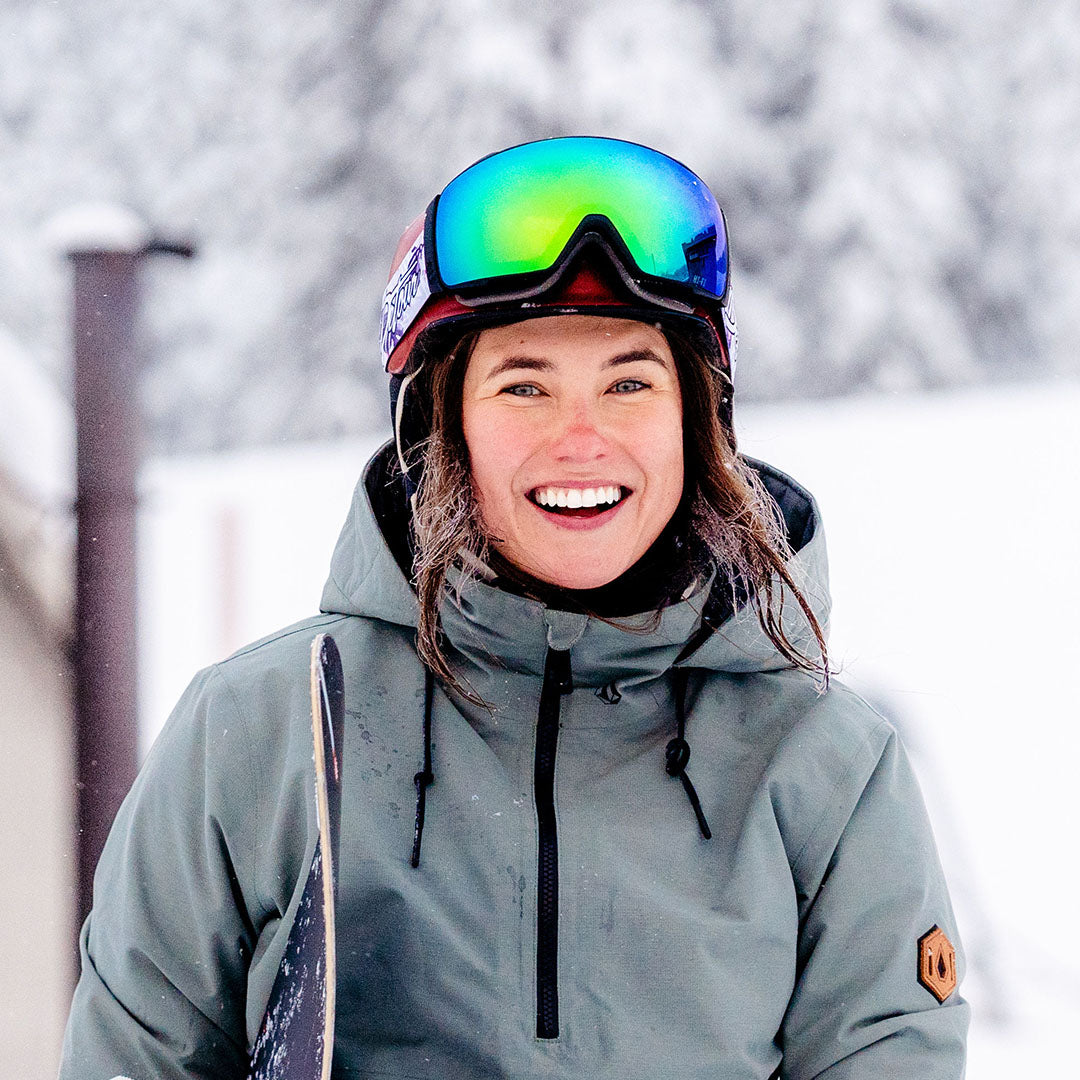 Man wearing mirrored goggles and gray beanie, holding skis in a snowy setting.